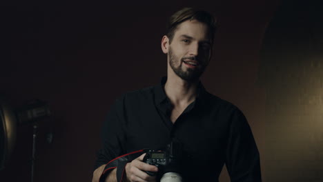 vista frontal de un joven fotógrafo caucásico tomando una foto con una cámara y sonriendo en el fondo oscuro del estudio