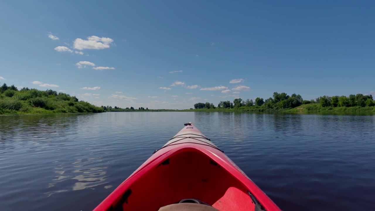 Prospettiva in prima persona di un kayak rosso che pagaia su un fiume tranquillo circondato da lussureggianti foreste verdi sotto un cielo estivo azzurro brillante con nuvole in Lettonia