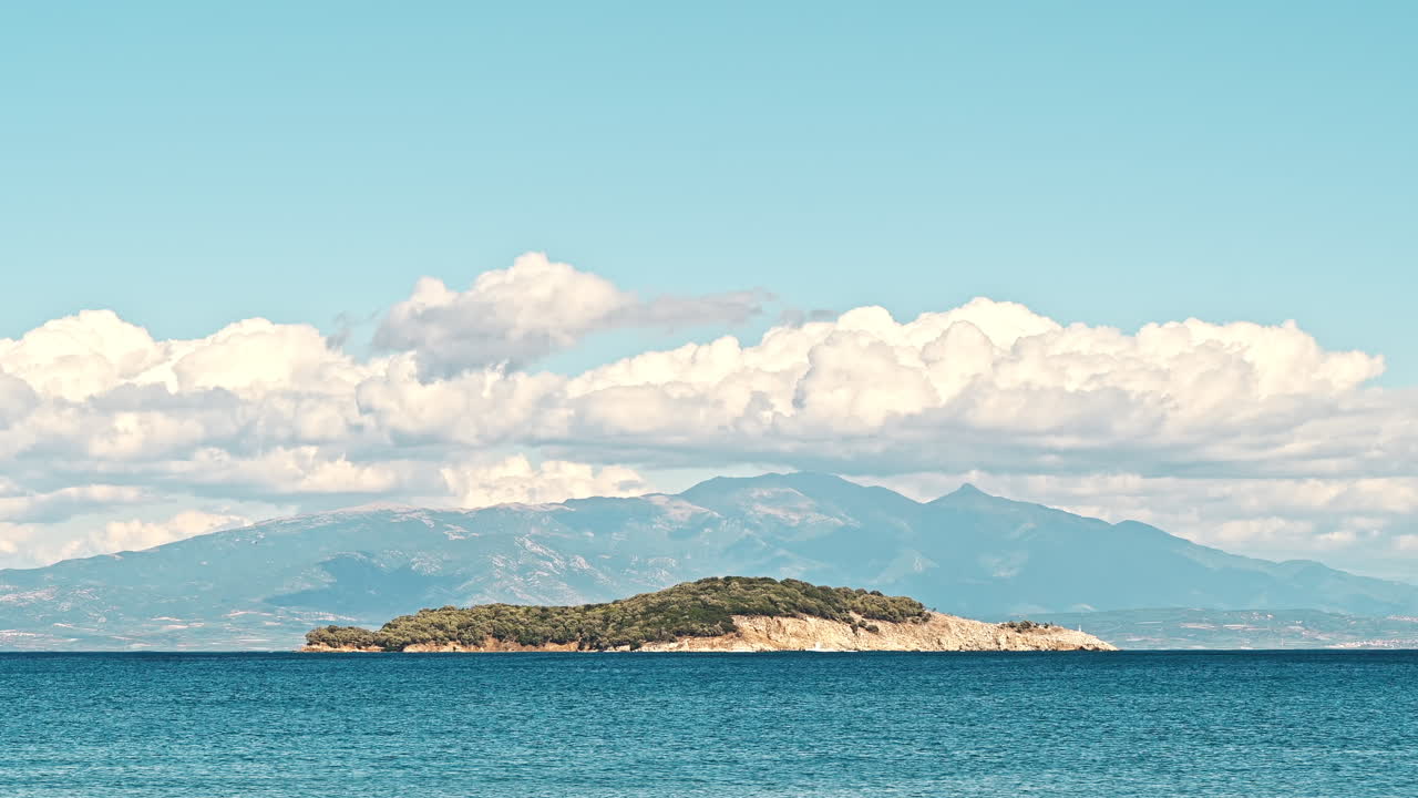 An island and a mountain reaching clouds in the distance with Aegean sea on the foreground in Greece