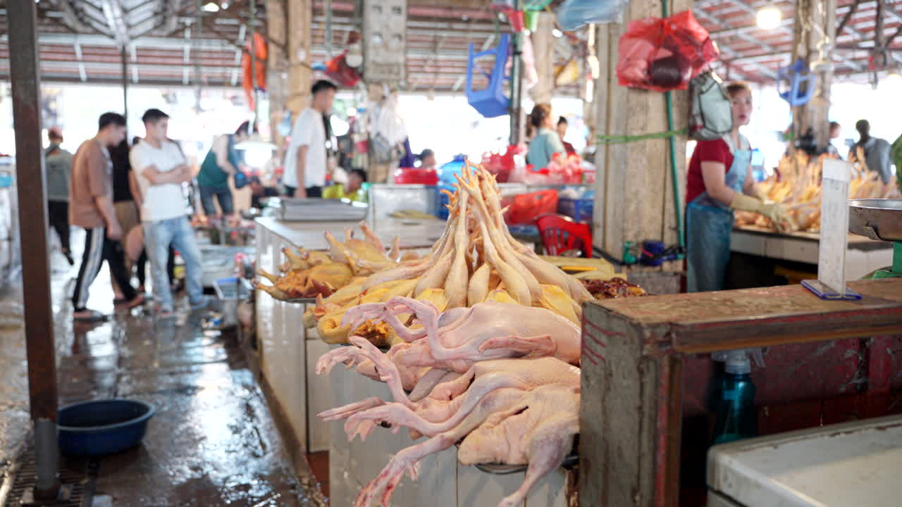 Busy wet market with meat vendors and fresh produce displayed, vibrant market life, fan sways above to keep flies off poultry as locals out of focus walk around