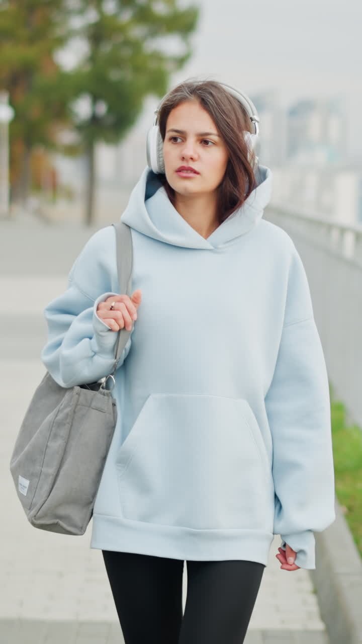 Giovane donna con una felpa con cappuccio blu che ascolta pensierosa la musica, sistemando la sua borsa mentre cammina lentamente lungo una ringhiera di ferro, godendosi una passeggiata all'aperto, un momento calmo e sereno nella natura.
