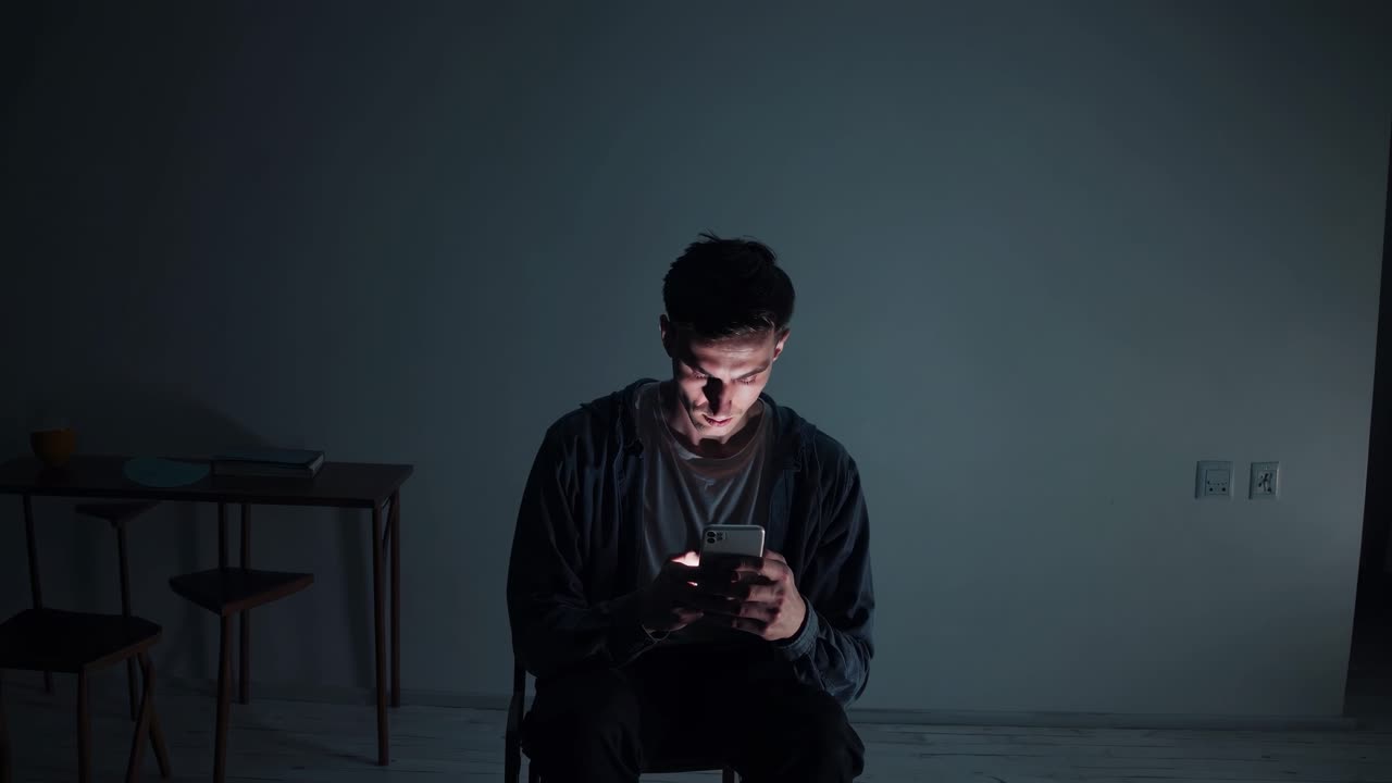 Young man using mobile phone sitting on a chair in a dark room ...