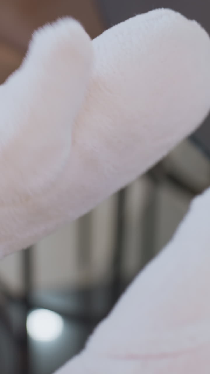 Close-up of hands in soft, plush winter gloves clapping together, the cozy fabric texture contrasts with the blurred background featuring a table and chair