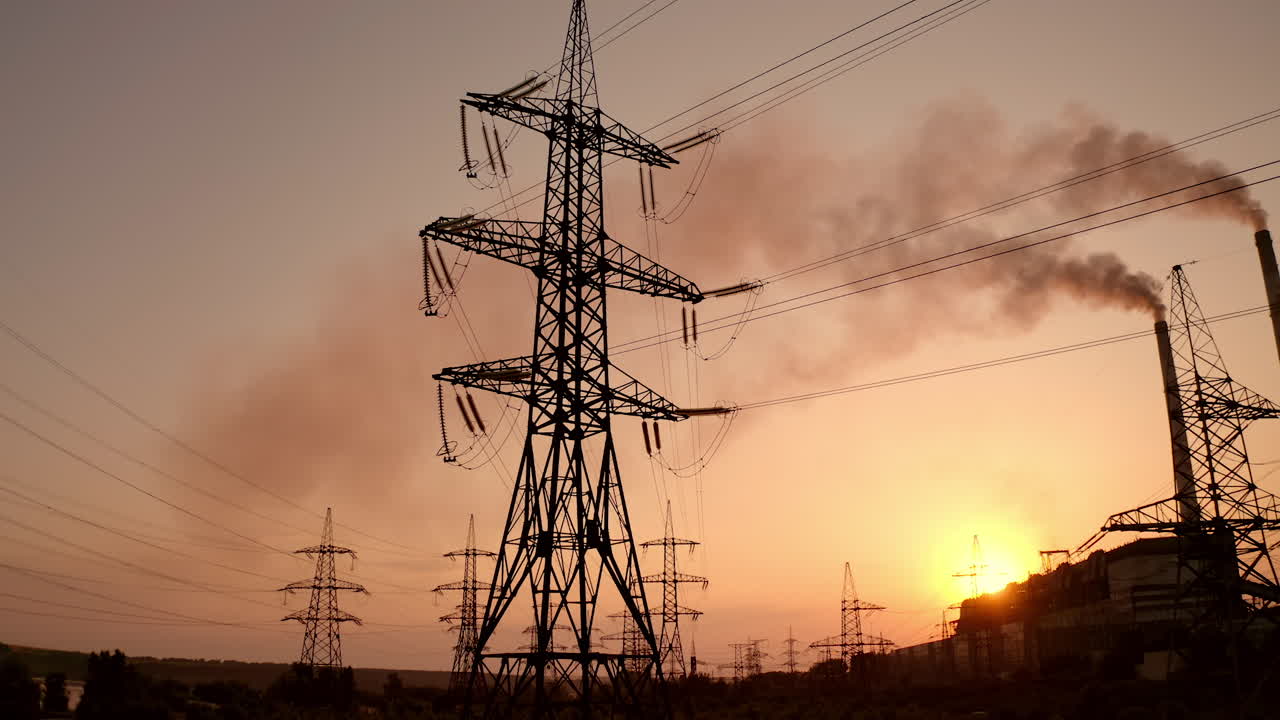Transmission lines near smoking industrial pipe at sunset. High-voltage electric power line and dark smoke released from factory in the evening