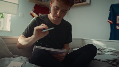 tilt up of caucasian teenage boy sitting on bed and making notes.