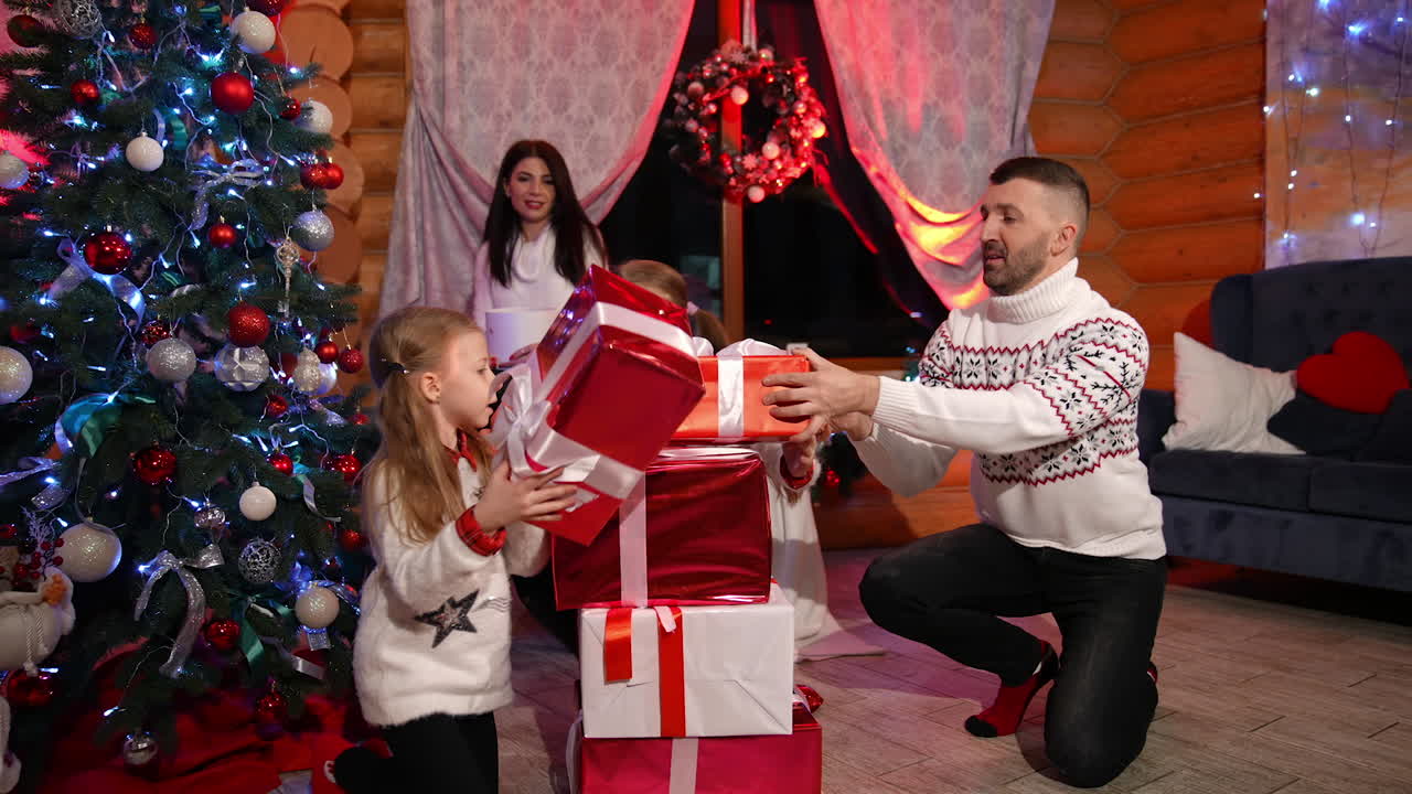 Happy family together at Christmas. Parents and their little daughters in warm white sweaters play with gift boxes near Christmas tree in the living-room.