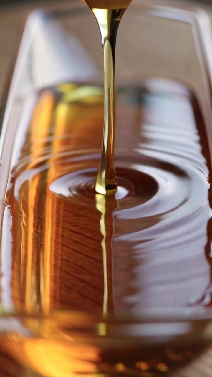 Video verticale: Versamento di sciroppo dorato da un mestolo, il flusso forma increspature in una ciotola di vetro per riempire una pozza.