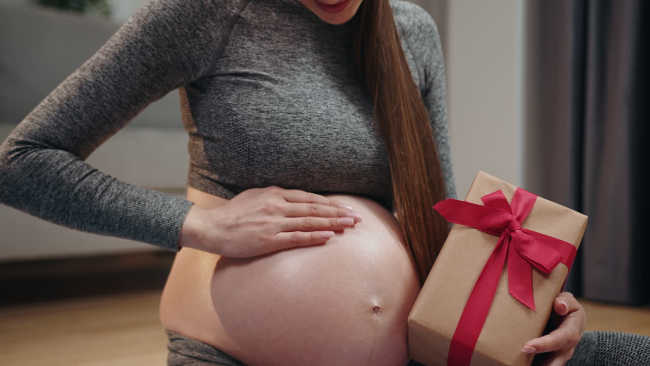 Pregnant woman in grey clothing cradles her belly while holding a brown gift box tied with a red
