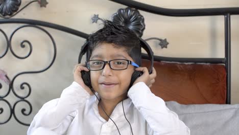 Lindo-Niño-Indio-Asiático-Caucásico-Con-Anteojos-Escuchando-Música-En-Los-Auriculares-Con-Las-Manos-En-La-Oreja-Sentado-En-La-Sala-De-Estar-Mirando-La-Vista-Frontal-De-La-Cámara