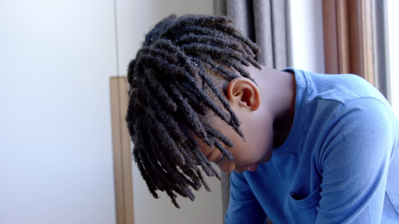Sad african american boy sitting next to window at home, slow motion