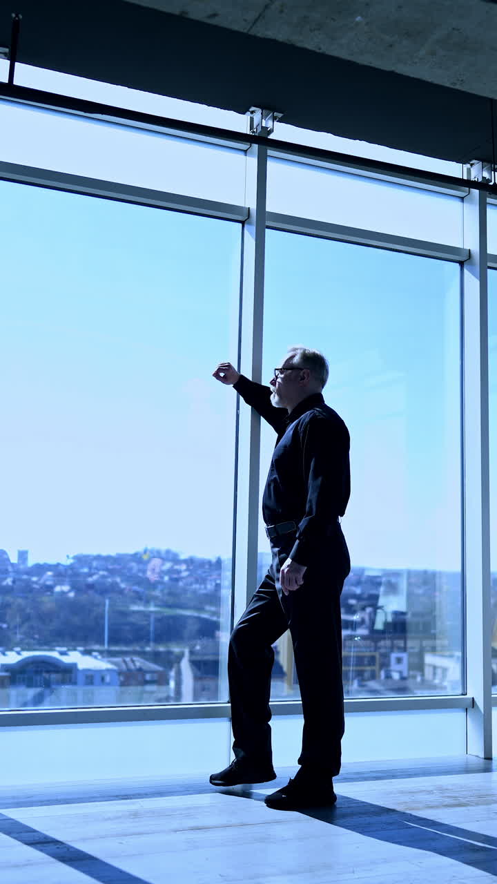 Old man wearing black shirt and trousers stands at the window. Man looks at city view thinking over something. Vertical video.