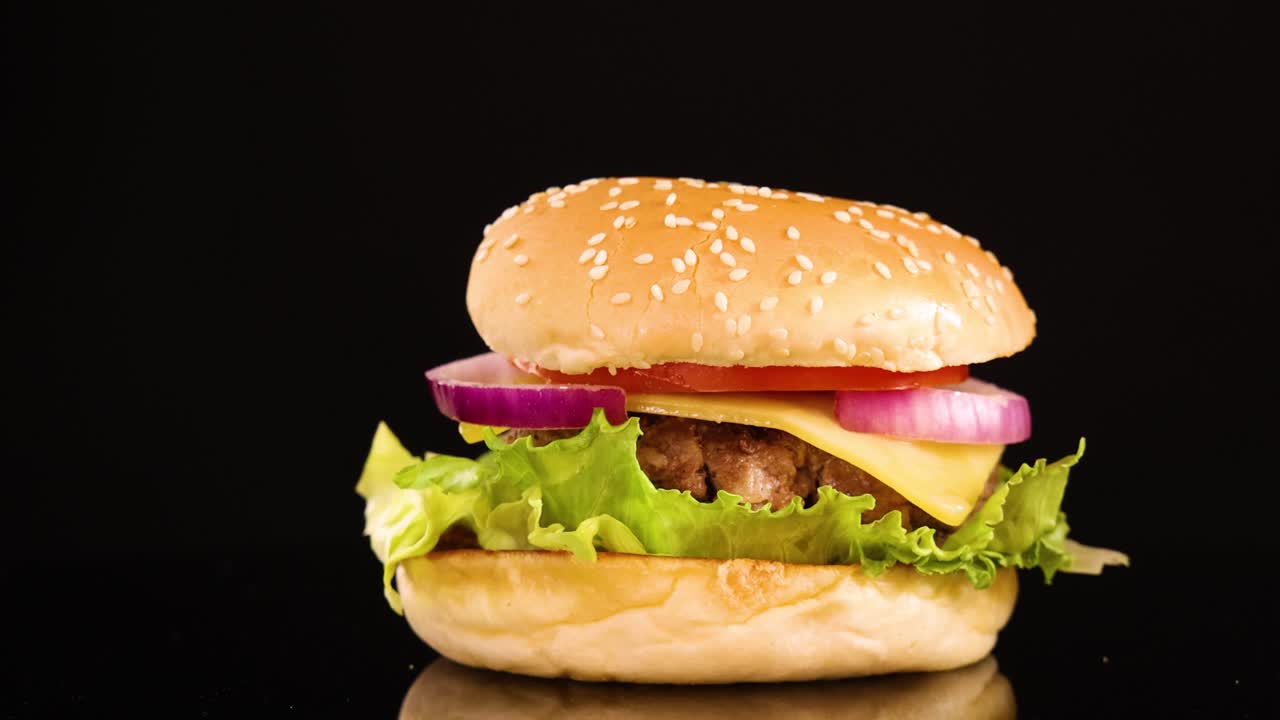 A beef burger with lettuce, onion, and tomato rotates smoothly on a reflective surface under bright studio lighting, isolated against a black background