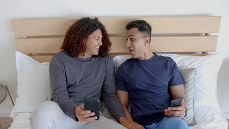 happy diverse gay male couple sitting on bed using smartphones, talking in the morning, slow motion