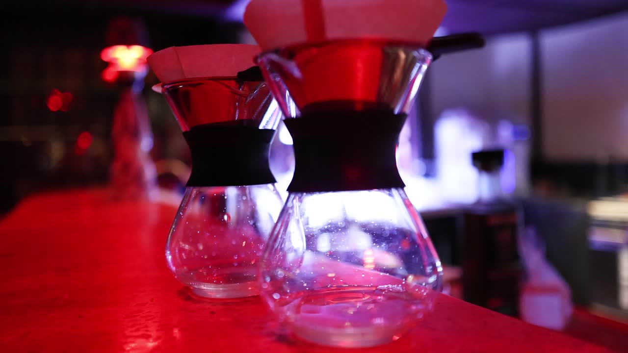 Two glass coffee drippers sit on a red bar counter with dramatic neon lighting effects