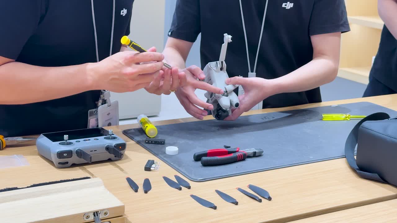 Two technicians assemble drone components on workbench under bright lighting, using tools and organized layout