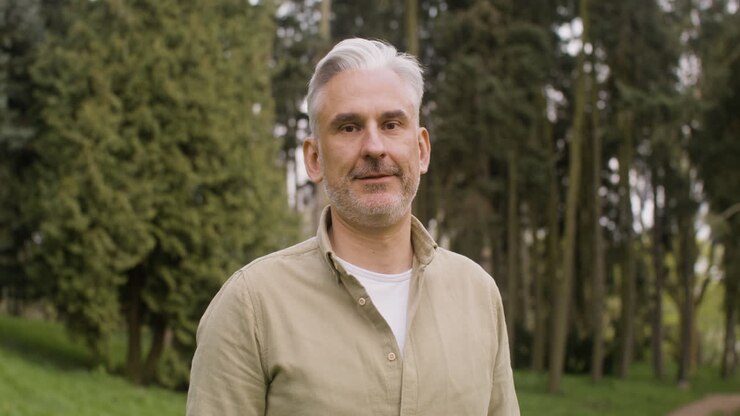 Ritratto di un uomo con i capelli grigi, in piedi nel parco e che guarda l'obiettivo.
