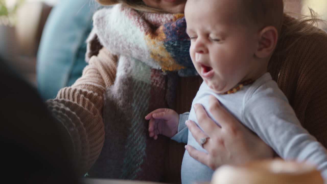 아기를 데리고 카페에 있는 젊은 엄마는 분주한 레스토랑에서 스마트폰으로 커피를 마시며 모성을 즐기고 있습니다.