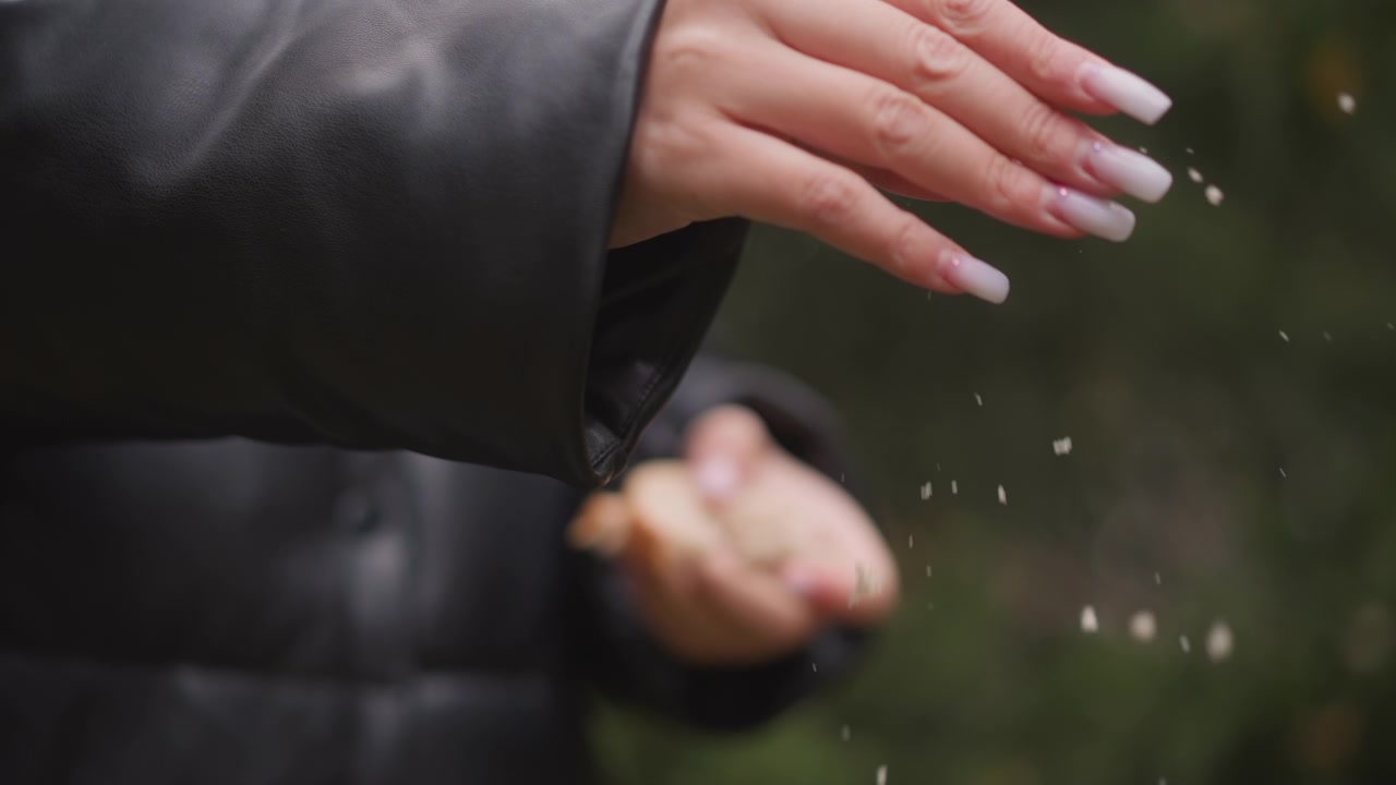 Primo piano di una donna che indossa una giacca di pelle nera, che tiene e spezza piccoli pezzi di cibo, con smalto bianco visibile. Lo sfondo è una vegetazione leggermente sfocata che suggerisce un ambiente esterno