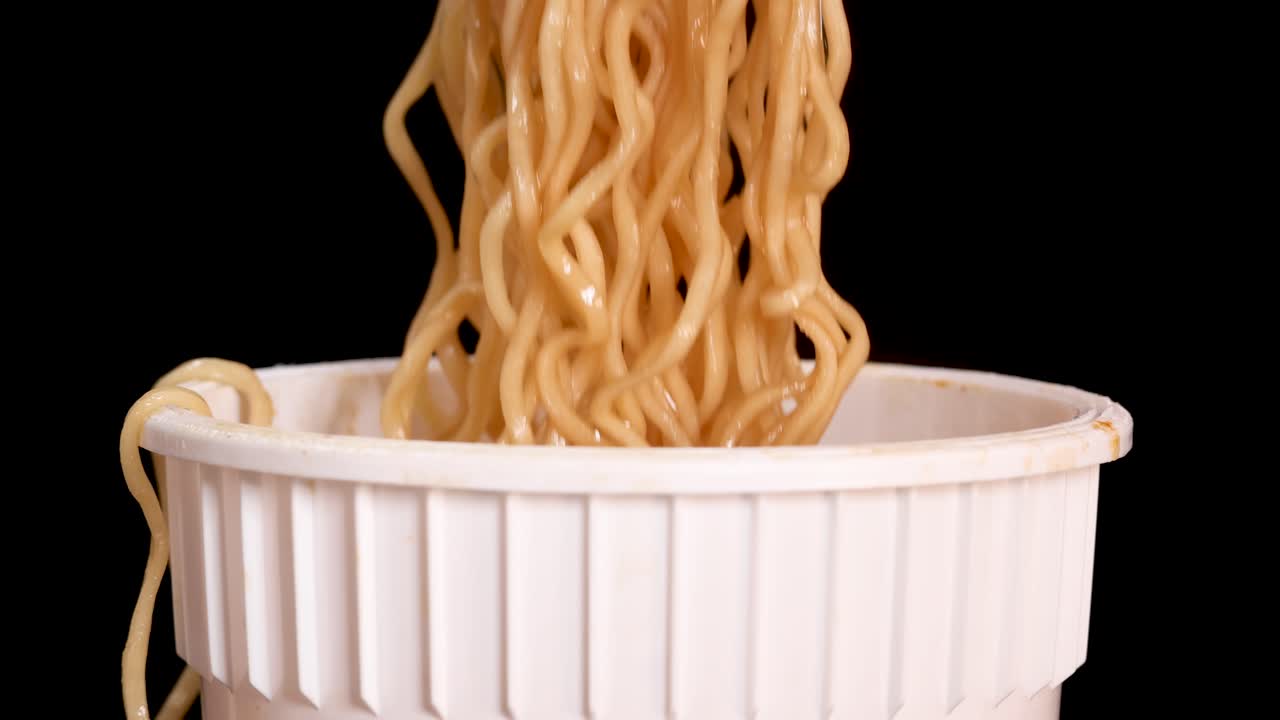 A fork lifts cooked instant noodles from a white disposable cup against a solid black background, with even studio lighting and steady close-up framing