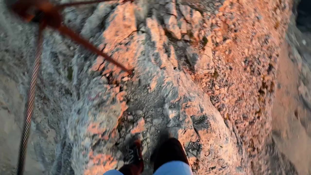 Caminhante atravessa cuidadosamente uma crista rochosa estreita com quedas íngremes em ambos os lados, segurando um cabo de segurança durante uma desafiadora rota de montanha em alta altitude.