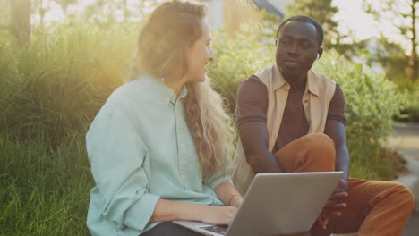 Multiethnic-Man-and-Woman-Sitting-with-Laptop-on-Street-and-Speaking