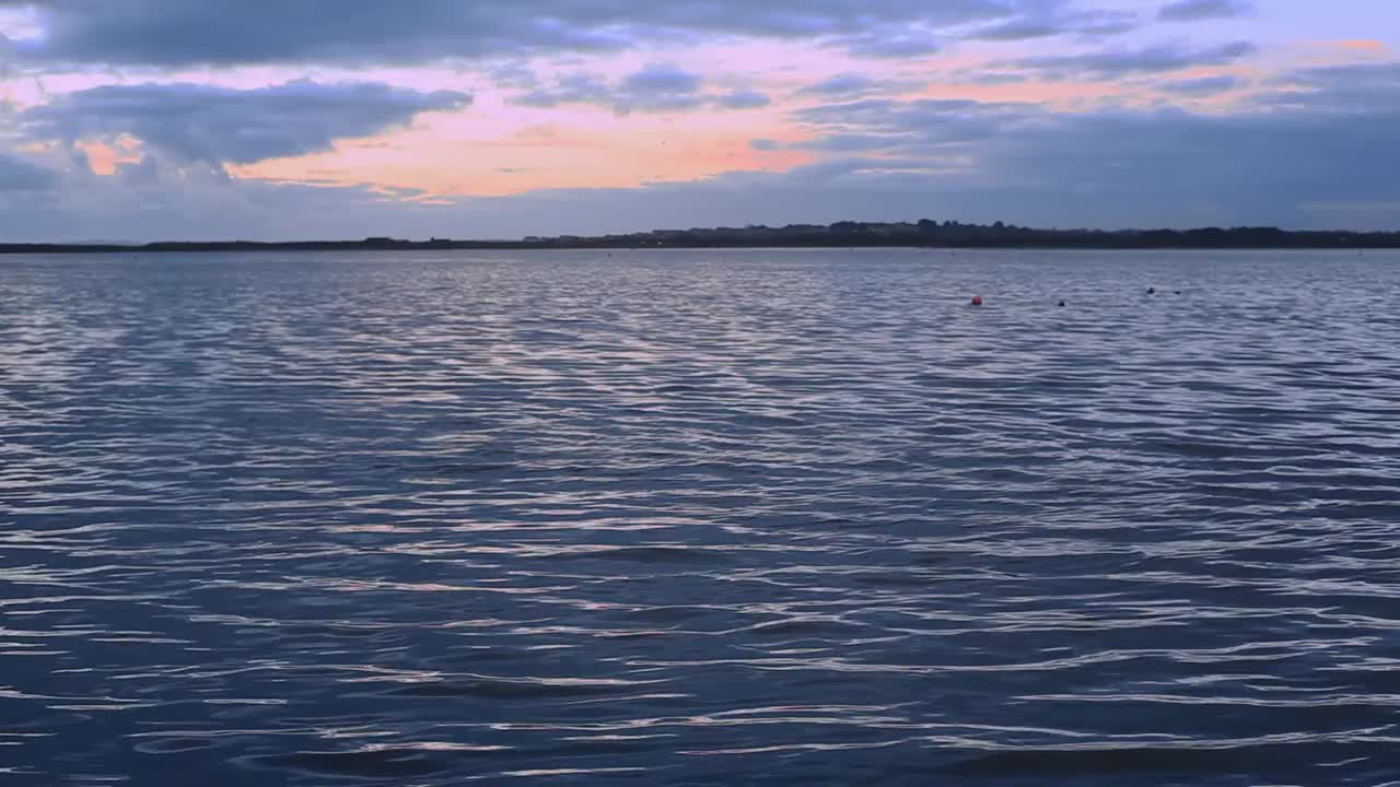 Bay waters sway peacefully; small ripples reflect raising or setting sun; water looks cool and calm. Blue sky with orange and yellow light patches. Camera is at a low angle. Footage in HD