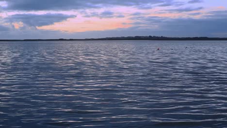 bay waters sway peacefully; small ripples reflect raising or setting sun; water looks cool and calm. blue sky with orange and yellow light patches. camera is at a low angle. footage in hd