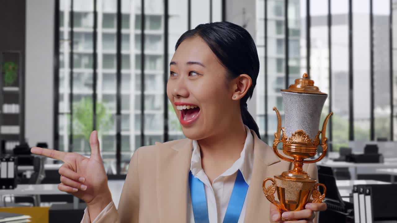 Close Up Of Asian Business Woman In A Suit With A Gold Medal Holding A Gold Trophy In Her Hands, Smiling, And Pointing To Side In The Office