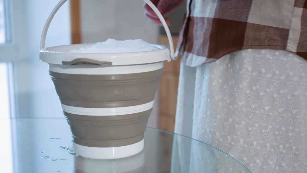 Serene figure carrying bucket full of snow places bucket gently on glass table in cozy indoor setting with wooden backdrop highlighting soft light and mindful seasonal ritual