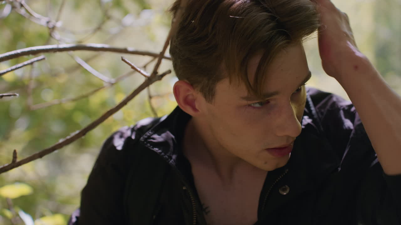 Young man with looks down thoughtfully while adjusting hair, standing under soft forest sunlight among branches, contemplating path through woodland with focused expression and subtle