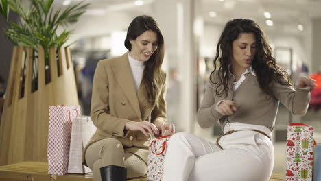 two women talking and interacting with shopping bags in a mall