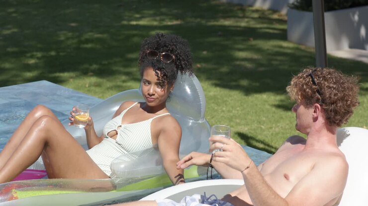 Coppia mista sdraiata su un galleggiante da piscina, la donna che si protende schizzando l'uomo, che ride flirtando con una bevanda.