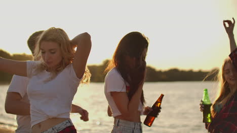 three female students with perfect bodies are dancing in short t-shirts with beer on the beach party at sunset. their long hair is flying on the wind.