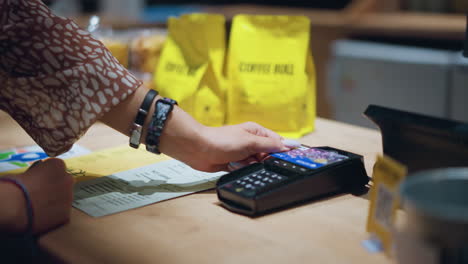 close-up of person in vintage outfit holding card over a card reader to make payment, with wrist adorned with bracelets and a watch, yellow coffee bags and menu papers are visible on table