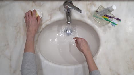 Bathroom-with-toothbrushes---old-woman's-hand's-are-washed-on-tap-water-with-soap,-then-rinsed-and-wiped-on-a-white-cloth-towel---top-view-static-close-up-shot-4k