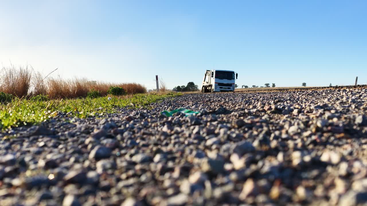 A heavy-duty truck travels along a natural gravel road in the Australian countryside, captured from a low, ground-level perspective in bright daylight