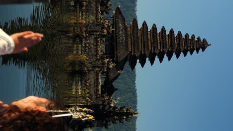 vertical ascending slow motion shot of a young traveler sitting on the shore danau batur in front of the water temple pura ulun danu temple overlooking the beautiful lake with beautiful nature