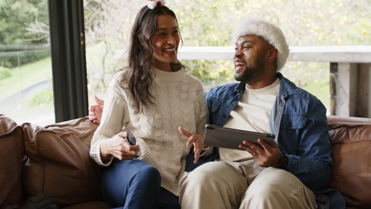 Diverse couple at home adjusting festive headgear, showing credit card and tapping tablet to shop