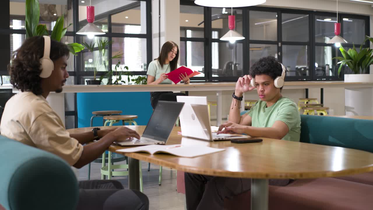 Uomini afroamericani che iniziano a studiare, digitando sui laptop, prendendo appunti e indossando cuffie in una lounge.