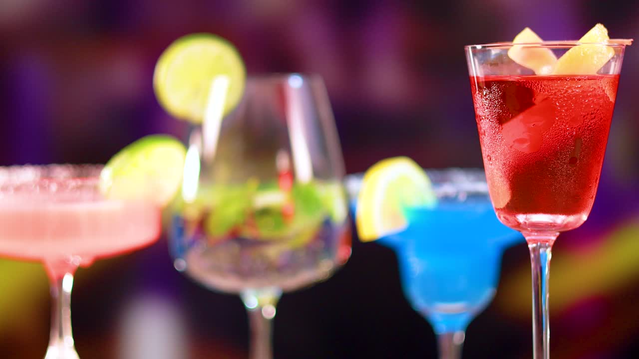 A series of cocktails in various glasses with vibrant colors and garnishes, set against a blurred, colorful background
