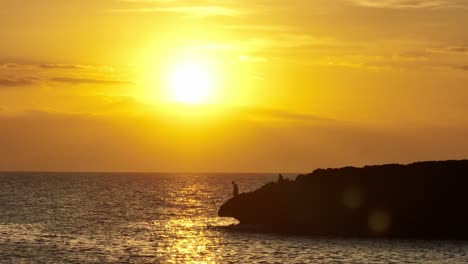 Contrasted-fishermen-shape-at-cliff-sea-shore-beach-with-sun-setting-below-golden-skyline