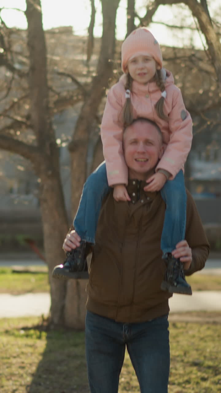 A man wearing a brown jacket carries a little girl in a pink cap and jacket on his neck as they walk through a sunlit park. The girl's legs dangle playfully
