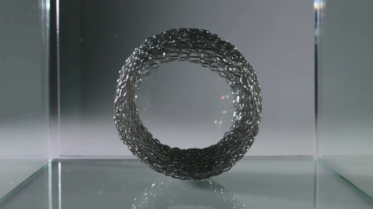 Starting metal mesh curling into torus inside clear glass tank on glossy base, forming stable ring