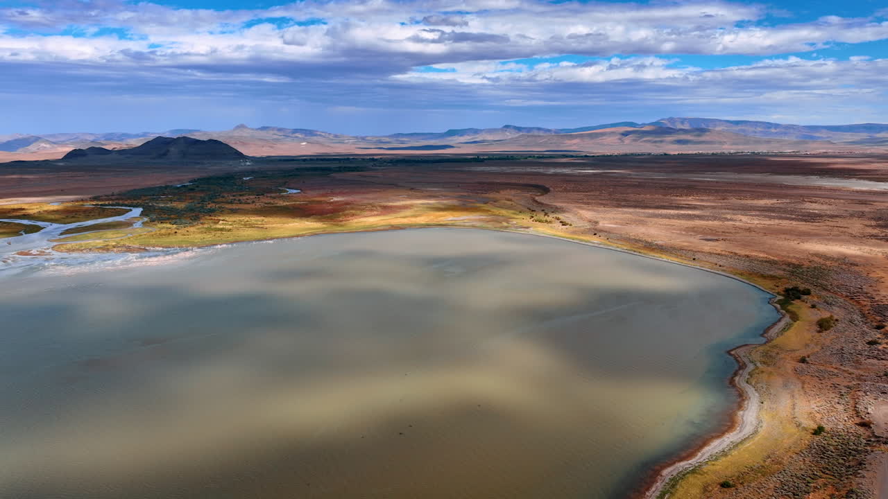 Flying over the lake with unclear water in the vast beautiful desert. Mountains with no vegetation at backdrop. Cumulus cloudscape covering the sky. Aerial view