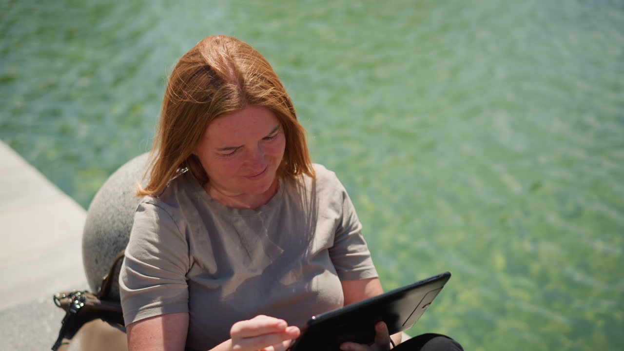 Student sitting near water operating tablet with gentle warmth, sliding through screen under sunlight, focused and relaxed mood, breeze moving hair, peaceful outdoor study