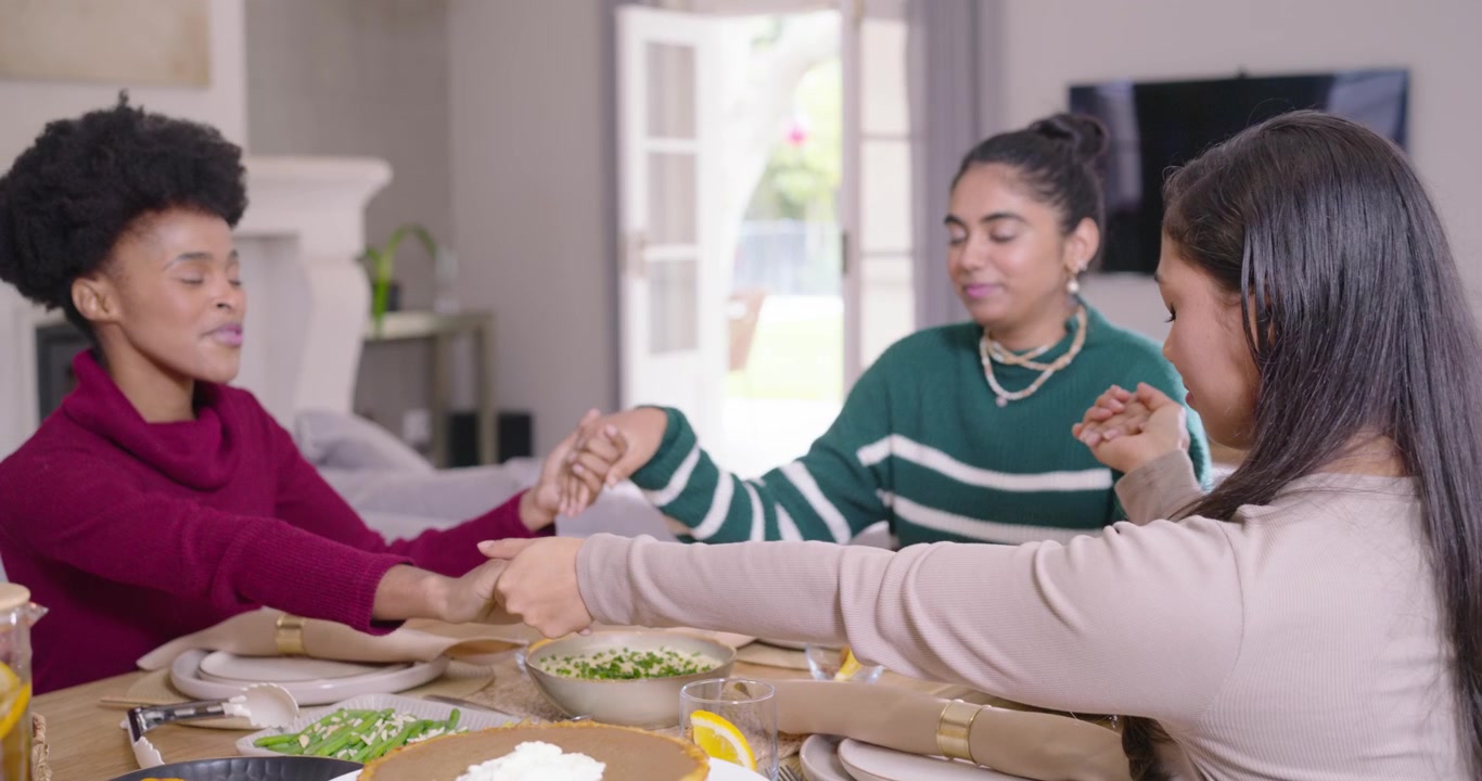 Amigas diversas colocando guardanapos, unindo as mãos e curvando as cabeças em gratidão à mesa de jantar