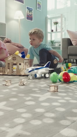 brother helps sister decorate roof of handmade wooden toy house. siblings sit on floor in nursery view through clearance in bunch of pencils in glass