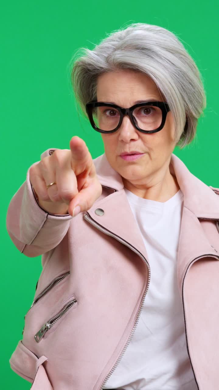 Donna matura con gli occhiali che indica direttamente la telecamera su schermo verde.