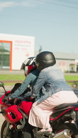 una mujer con una camisa a cuadros monta una bicicleta roja con su amiga sosteniéndola por detrás, ambos usan cascos, con árboles al lado, y un edificio lejano con vidrio