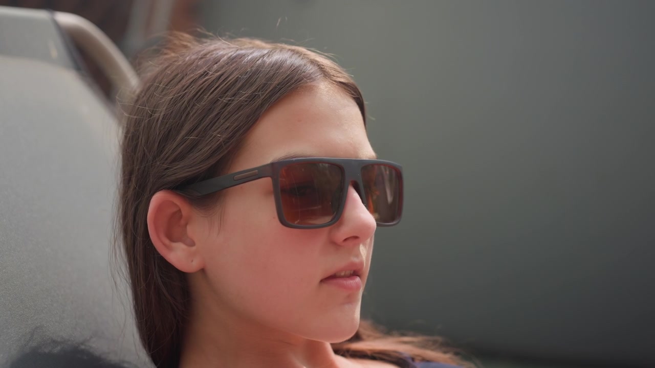 Young woman wearing sunglasses reclines in chair occasionally moving her arm. scene features soft natural light and outdoor background with muted colors